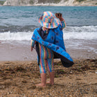 A child on a beach wearing a UV Protective Sun Hat Multi Stripe, holding a stone, with sea and cliffs in the background.